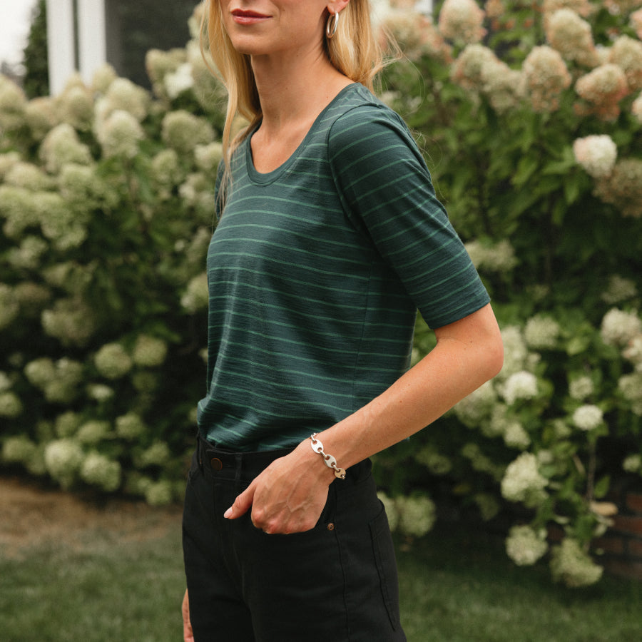 A woman wearing a dark green short-sleeve t-shirt with thin horizontal green stripes, paired with black pants, standing outdoors in front of a background of green bushes with white flowers.