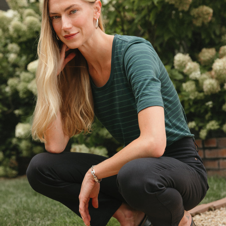 A woman crouching outdoors in a garden, wearing a dark green short-sleeve top with thin horizontal stripes and black pants.