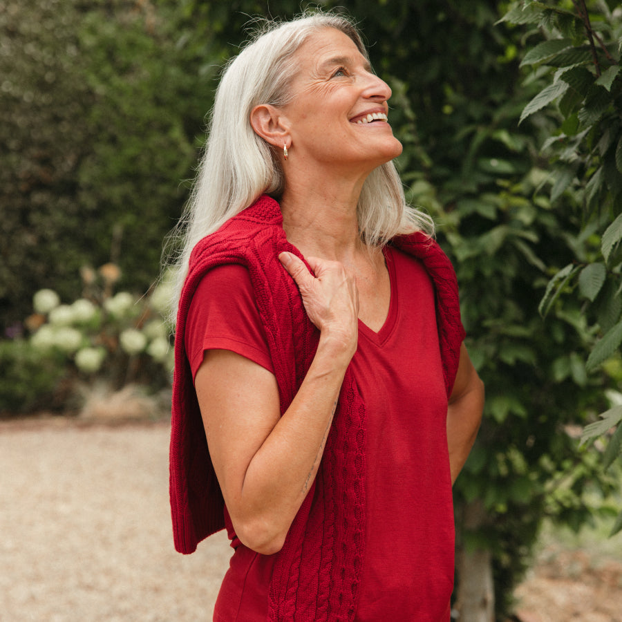 A smiling woman with long white hair stands outdoors wearing a red cable knit draped over her shoulders over a matching red top, with green foliage in the background.