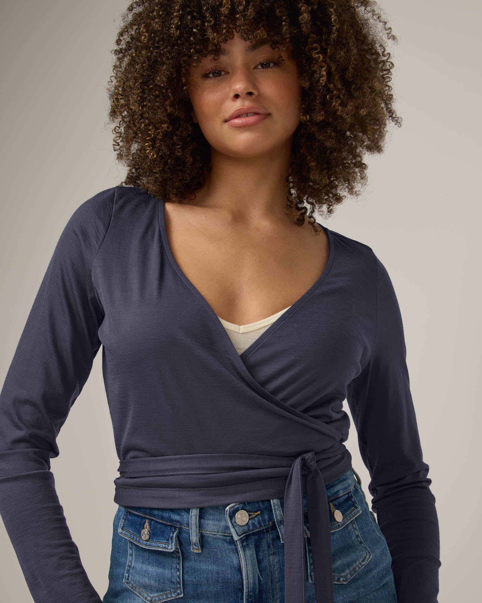 Model wearing Natalia Wrap Top in Odyssey Gray, a slate blue-gray color. Natalia Wrap Top is a merino wool long sleeve, v-neck, slim fit top available in sizes XS-3X.