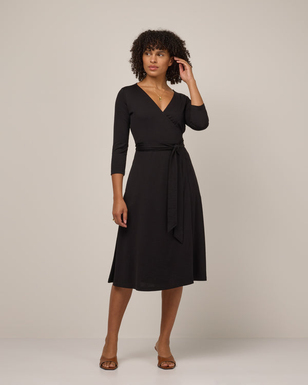 Model wearing Alana Wrap Dress in Black. Alana Wrap Dress is a merino wool long sleeve, v-neck, midi fit & flare dress with pockets available in sizes XS-3X.