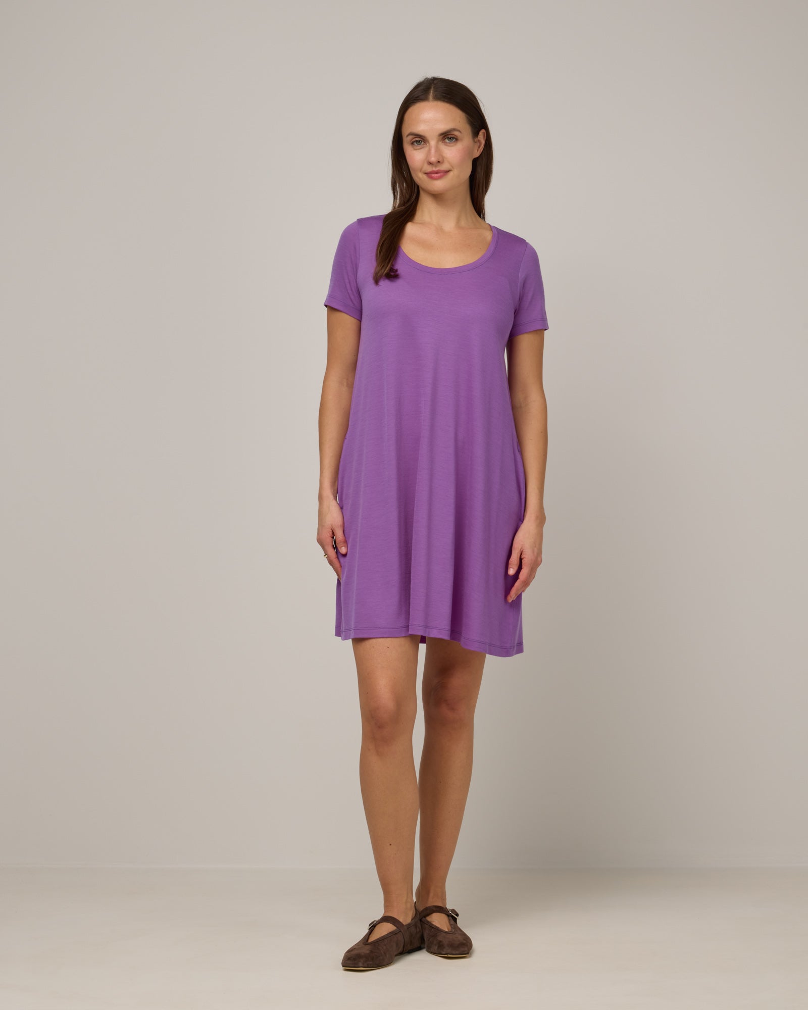 Model wearing Maggie Swing Dress in Purple Pansy, a bright, saturated purple color. Maggie Swing Dress is a merino wool short sleeve, scoop neck, knee length swing dress with pockets available in sizes XS-3X Regular, XS-3X Long.