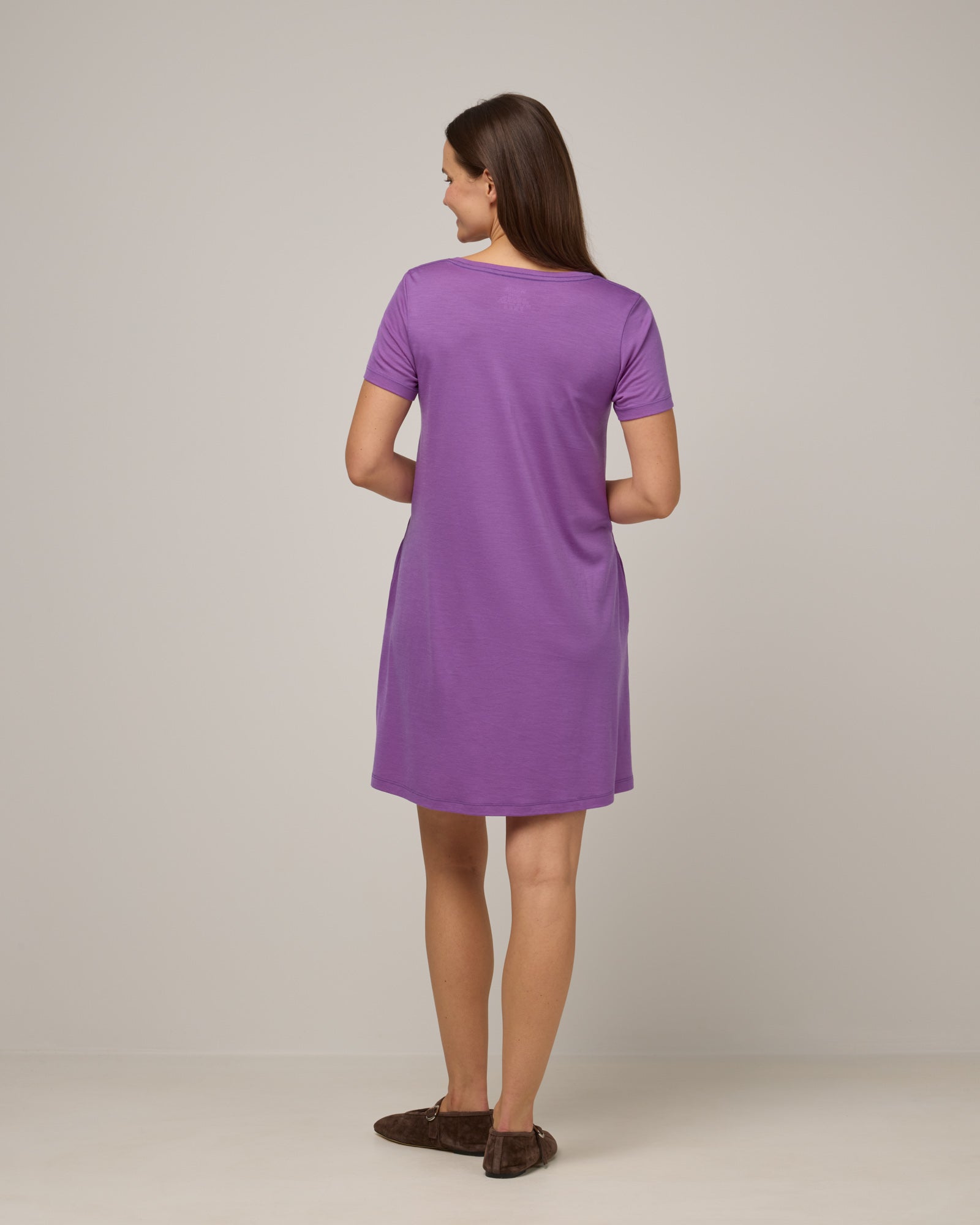 Model wearing Maggie Swing Dress in Purple Pansy, a bright, saturated purple color. Maggie Swing Dress is a merino wool short sleeve, scoop neck, knee length swing dress with pockets available in sizes XS-3X Regular, XS-3X Long.