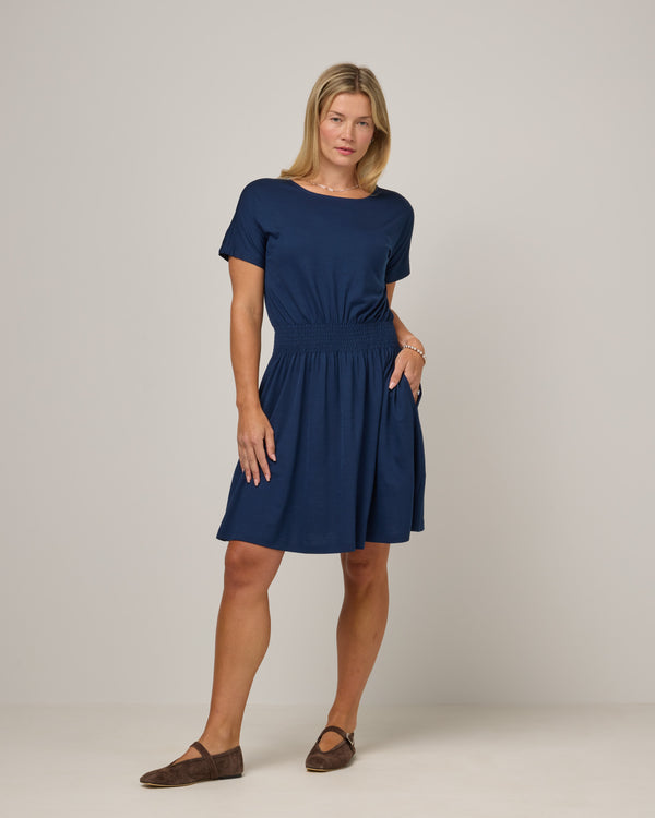 Model wearing Norma Smocked Waist Dress in Marine Blue, a deep royal blue color. Norma Smocked Waist Dress is a merino wool short sleeve, crew neck, knee length fit & flare dress with pockets available in sizes XS-3X.