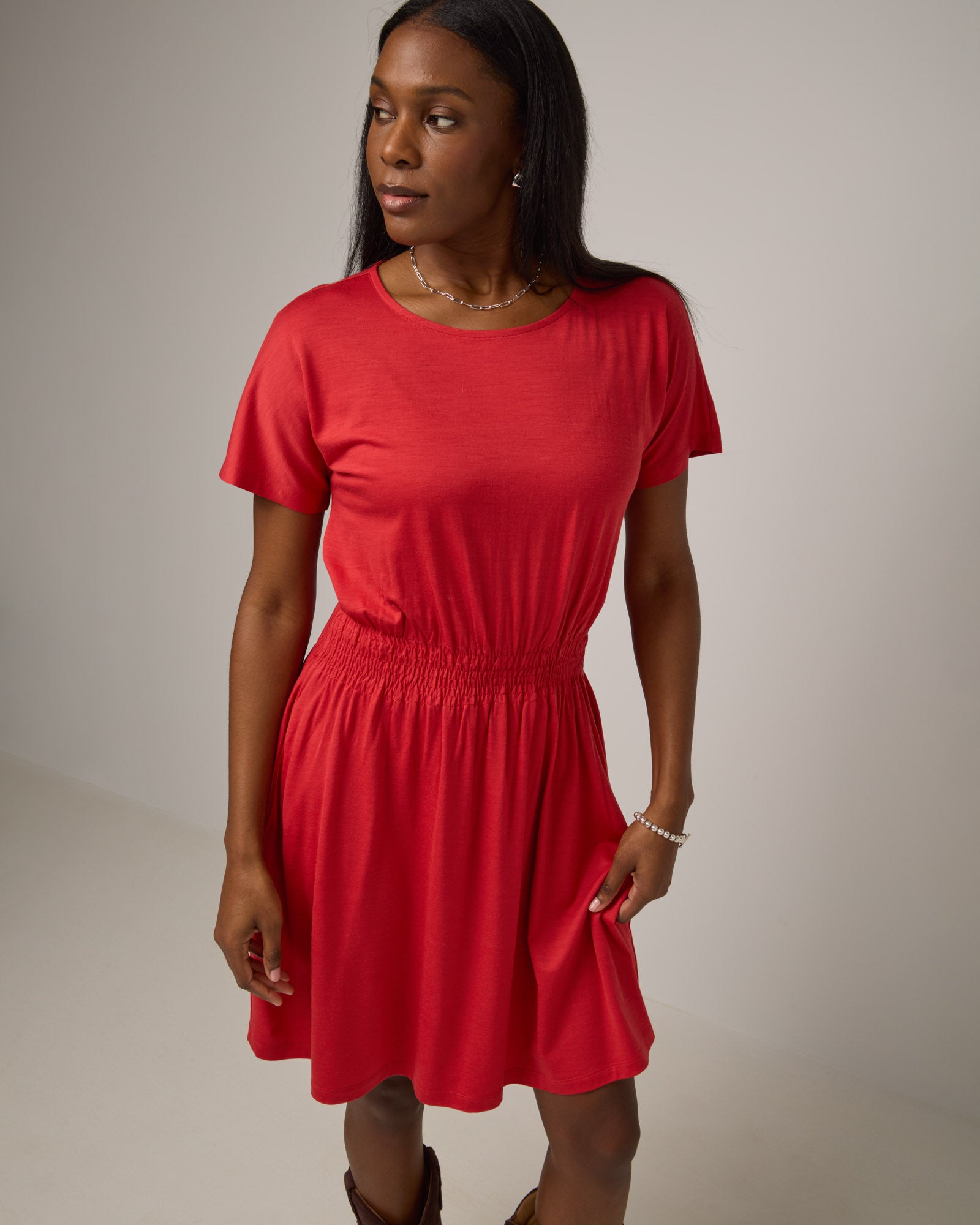 Model wearing Norma Smocked Waist Dress in Fruit Punch, a vivid red-orange color. Norma Smocked Waist Dress is a merino wool short sleeve, crew neck, knee length fit & flare dress with pockets available in sizes XS-3X.