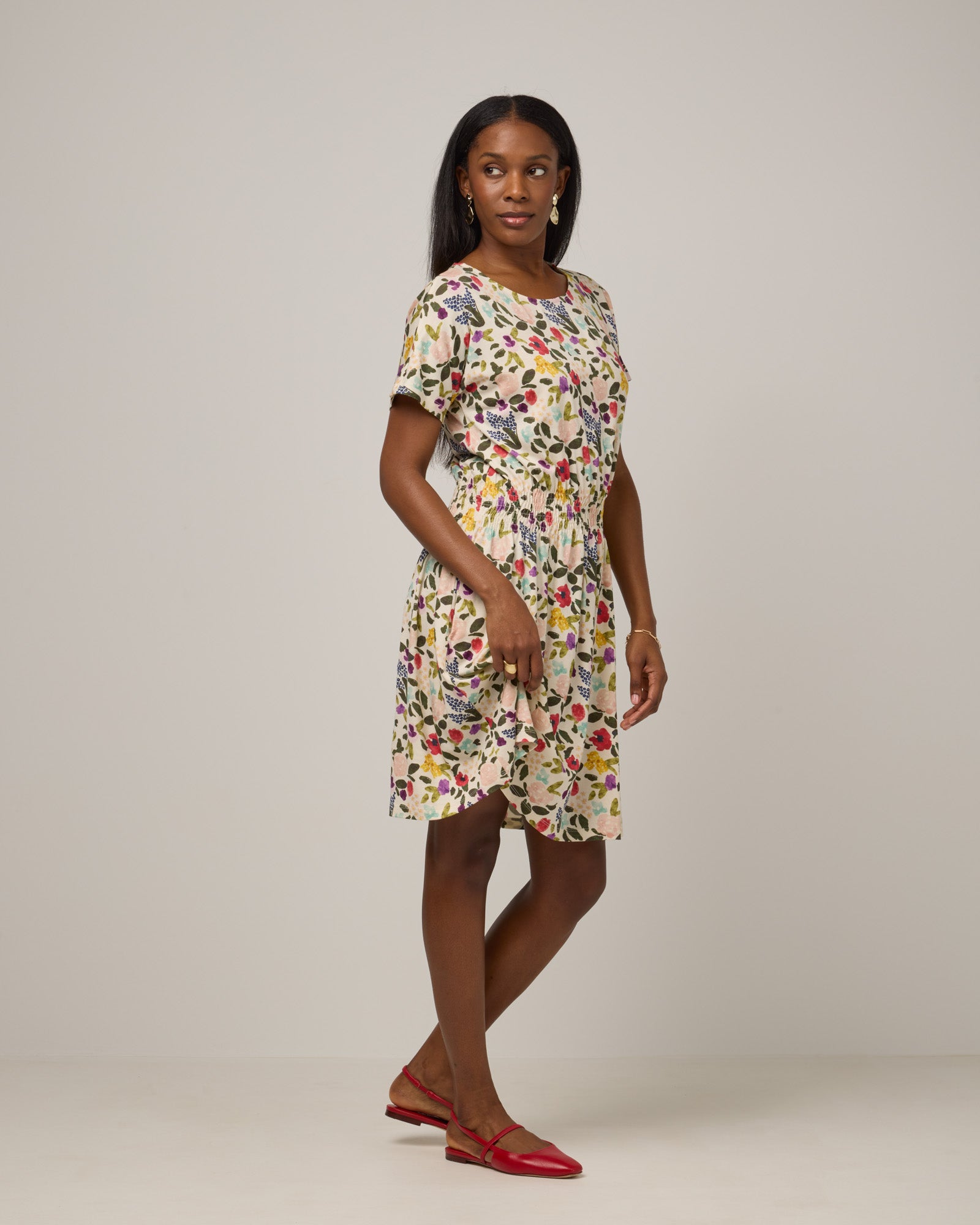Model wearing Norma Smocked Waist Dress in Watercolor Floral Print, a multi-colored floral print on a cream background. Norma Smocked Waist Dress is a merino wool short sleeve, crew neck, knee length fit & flare dress with pockets available in sizes XS-3X.