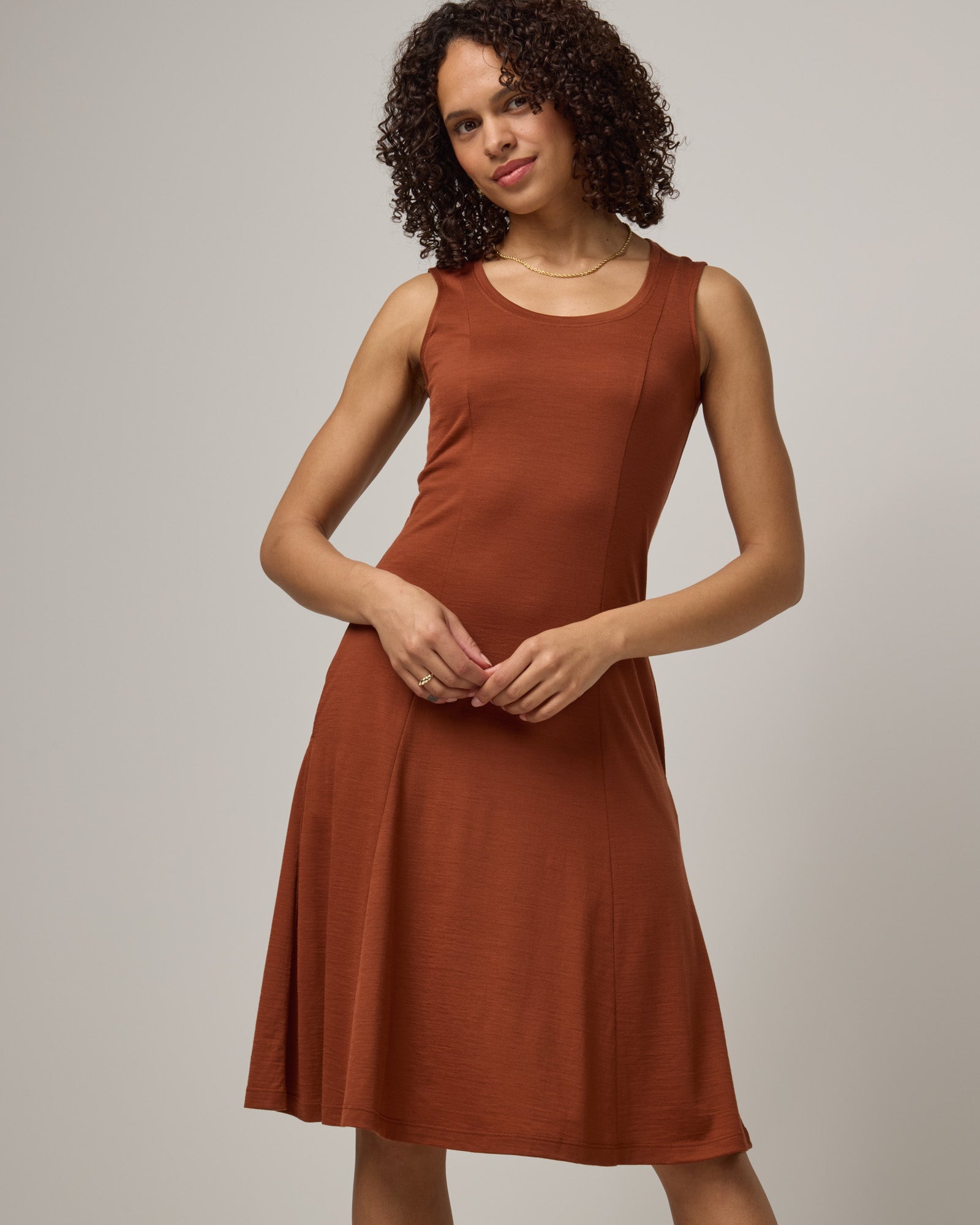 Model wearing Flora Swing Dress in Cinnamon, a reddish-brown rust color. Flora Swing Dress is a merino wool sleeveless, scoop neck, knee length swing dress with pockets available in sizes XS-3X.