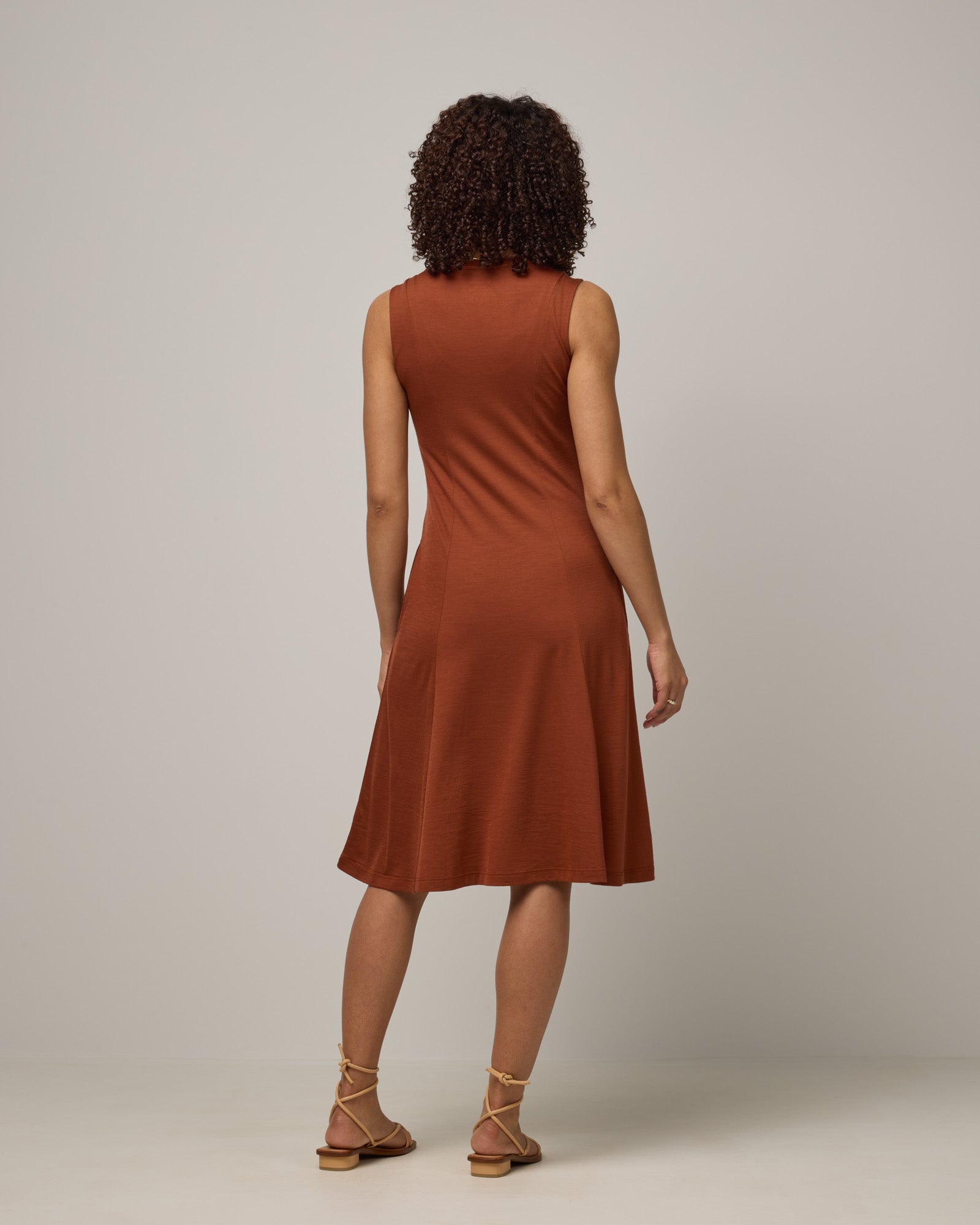 Model wearing Flora Swing Dress in Cinnamon, a reddish-brown rust color. Flora Swing Dress is a merino wool sleeveless, scoop neck, knee length swing dress with pockets available in sizes XS-3X.