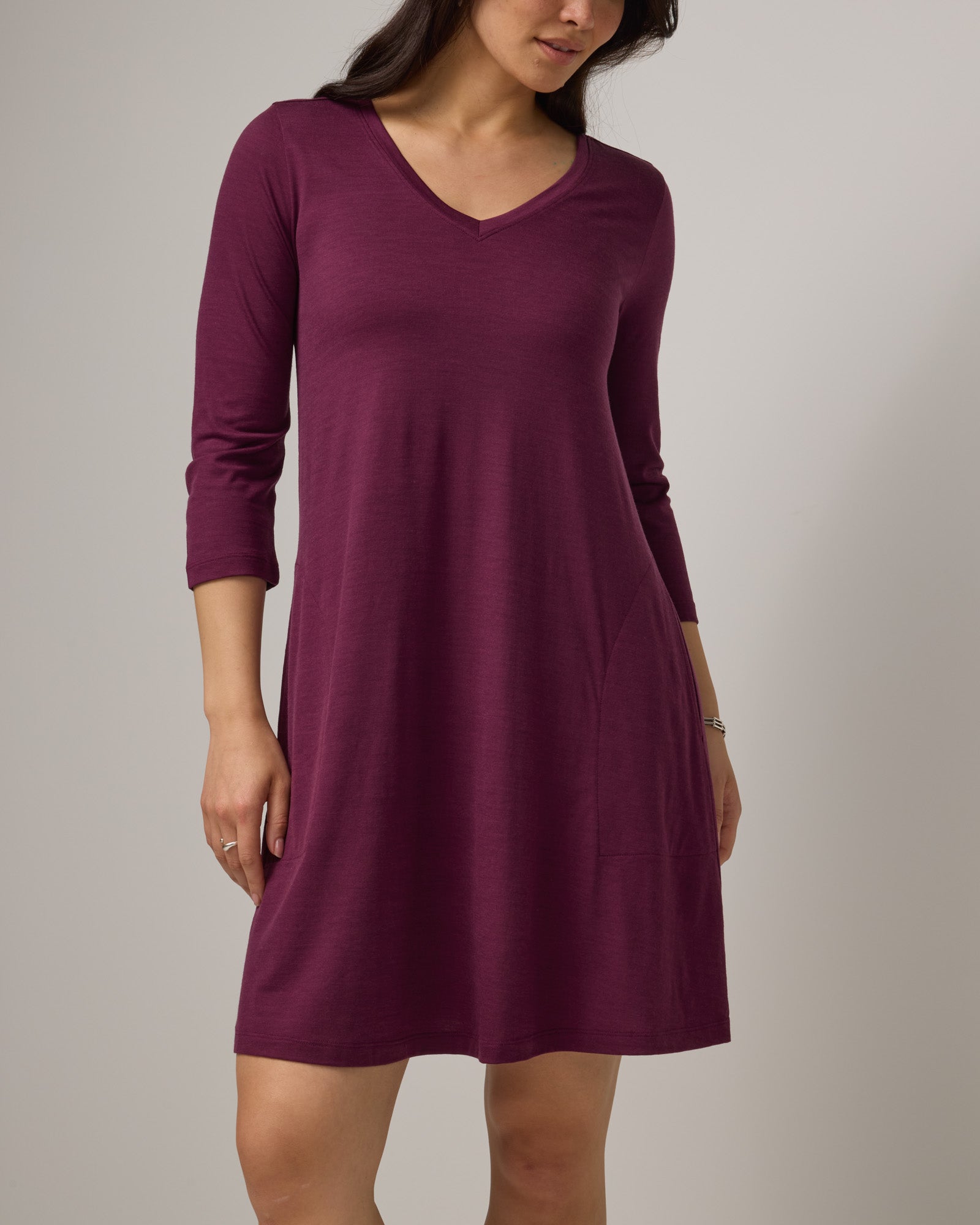 Model wearing Willow Swing Dress in Plum, a reddish-purple color. Willow Swing Dress is a merino wool long sleeve, v-neck, knee length swing dress with pockets available in sizes XS-3X Regular, XS-3X Long.