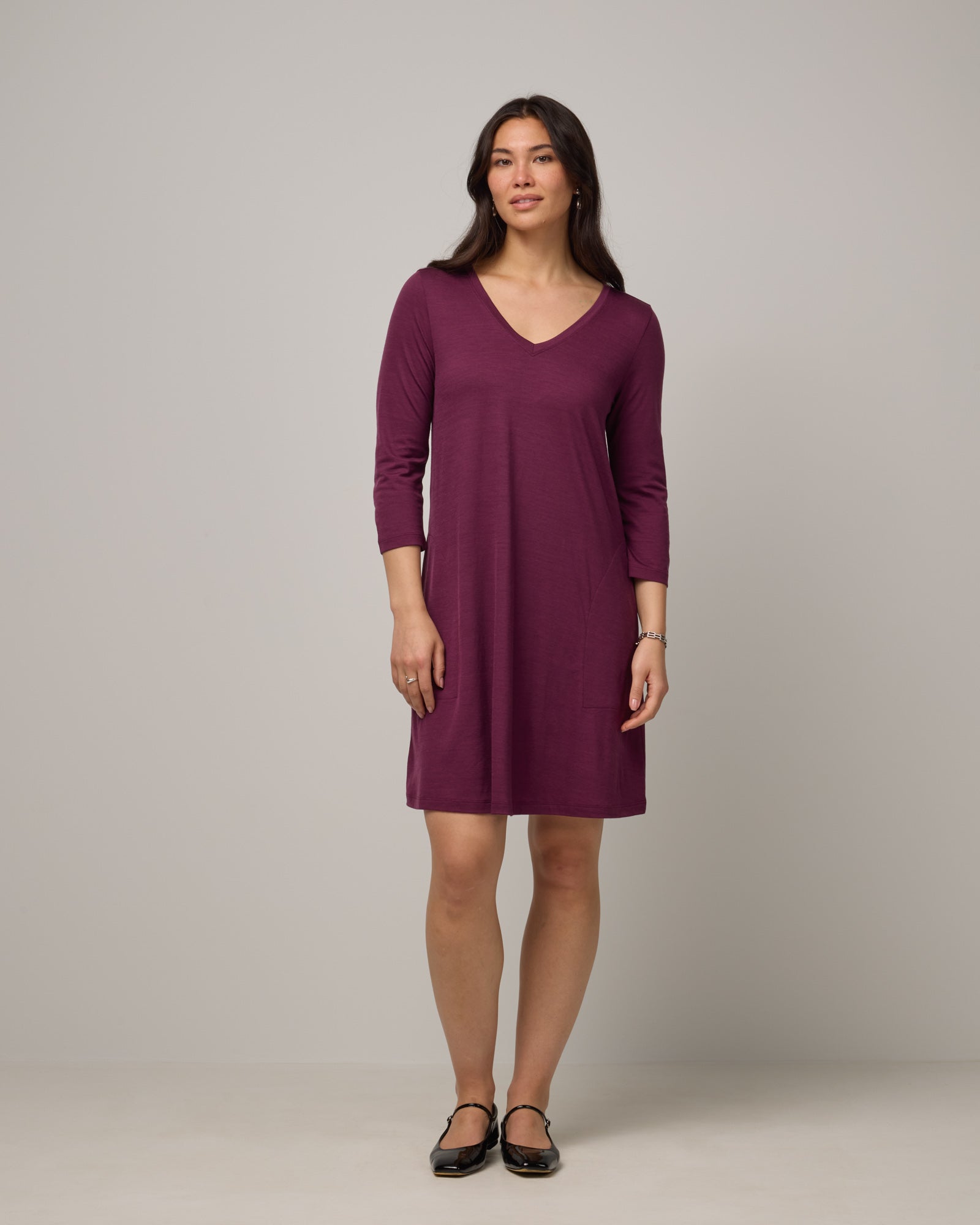 Model wearing Willow Swing Dress in Plum, a reddish-purple color. Willow Swing Dress is a merino wool long sleeve, v-neck, knee length swing dress with pockets available in sizes XS-3X Regular, XS-3X Long.