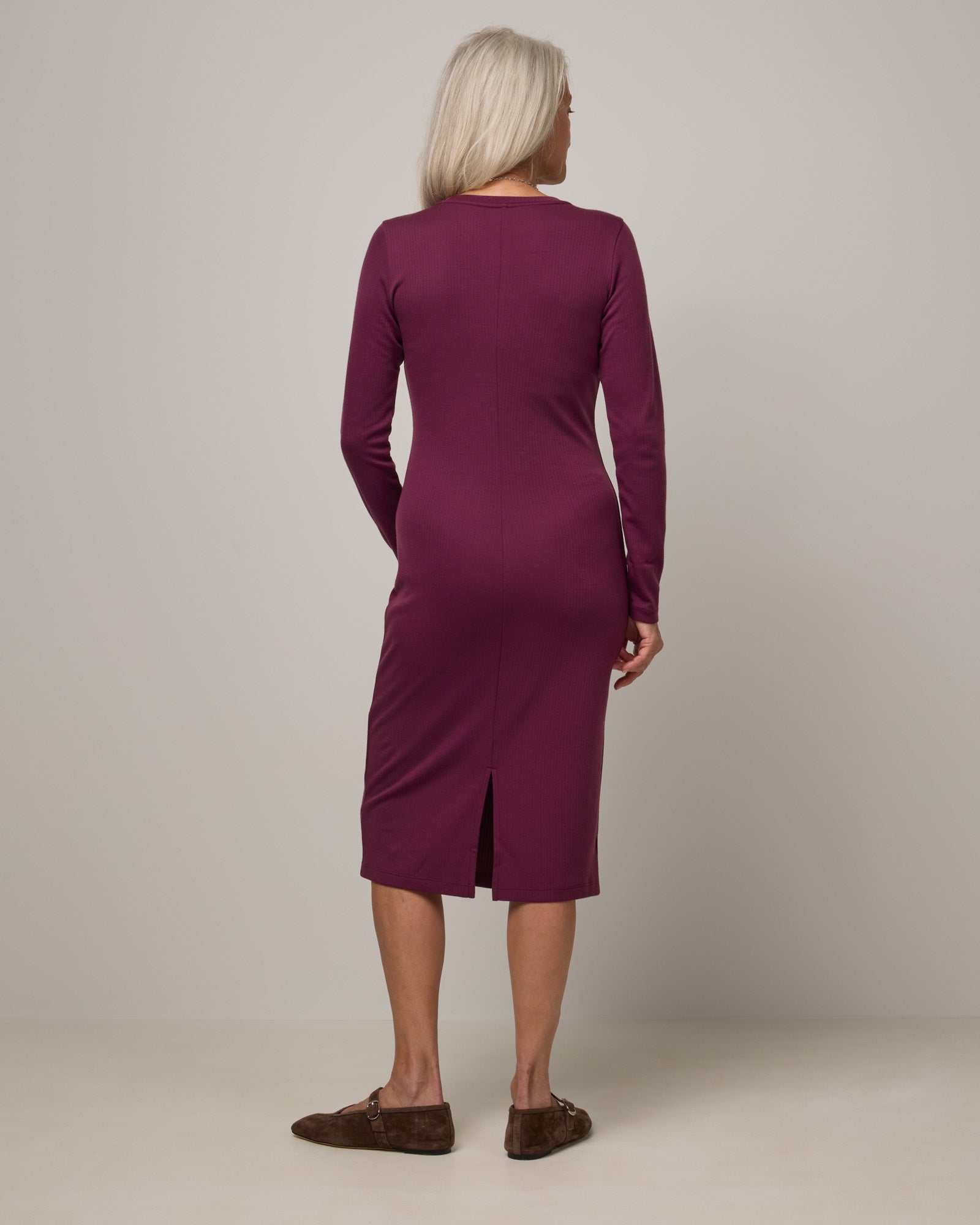 Model wearing Lex Ribbed Dress in Plum, a reddish-purple color. Lex Ribbed Dress is a merino wool long sleeve, scoop neck, fitted midi dress available in sizes XS-XXL Regular & XS-XL Petite.