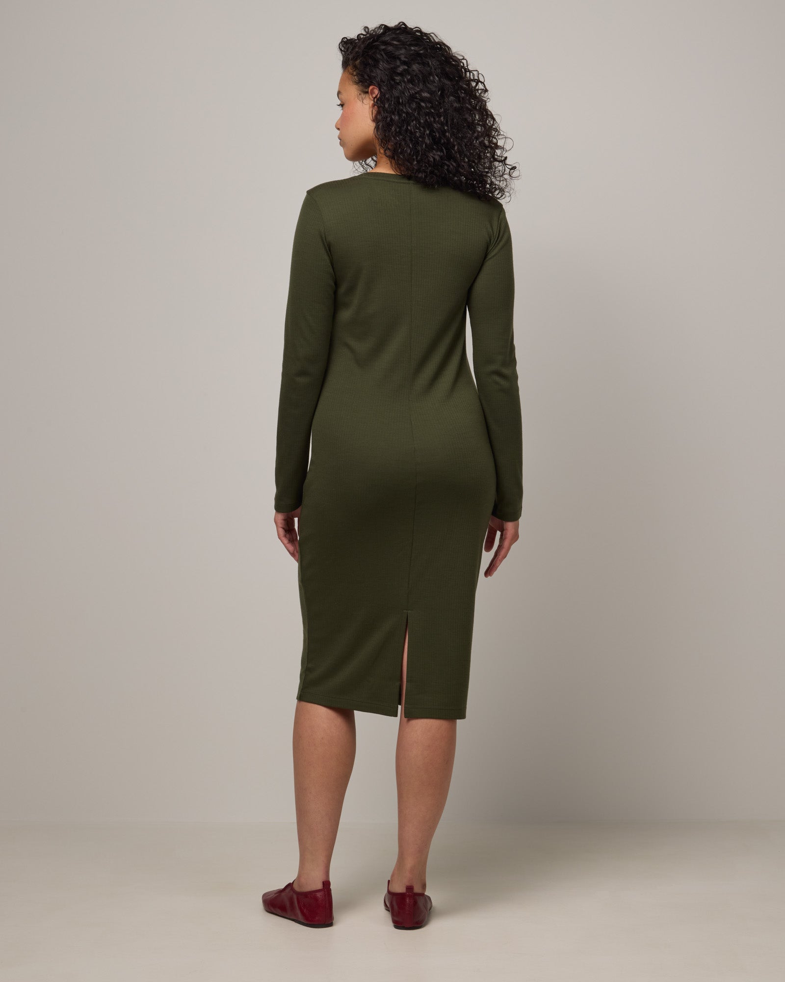 Model wearing Lex Ribbed Dress in Forest Night, a dark forest green color. Lex Ribbed Dress is a merino wool long sleeve, scoop neck, fitted midi dress available in sizes XS-XXL Regular & XS-XL Petite.