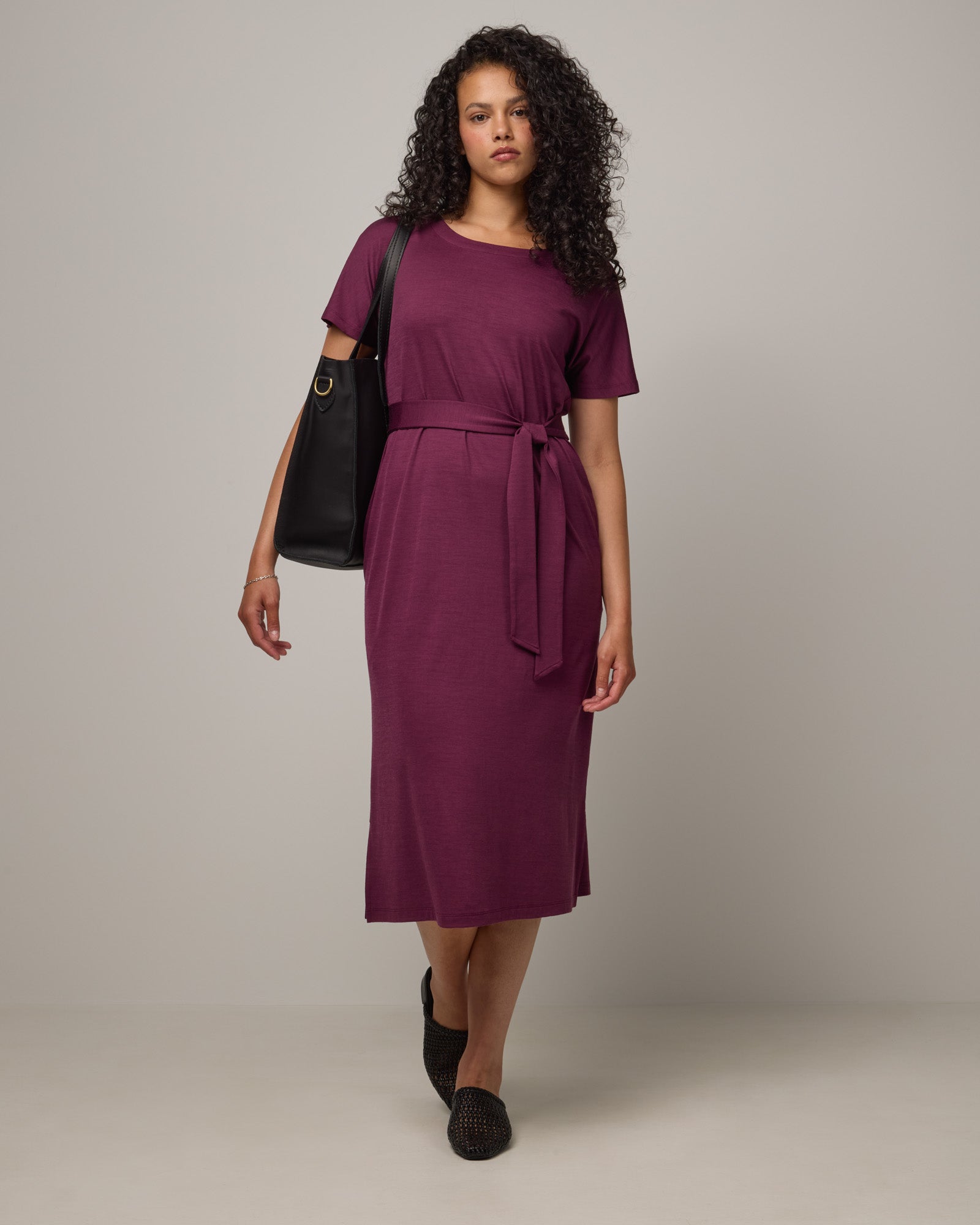 Model wearing Cora Shift Dress in Plum, a reddish-purple color. Cora Shift Dress is a merino wool short sleeve, midi shift dress with pockets and removable belt available in sizes XS-3X.