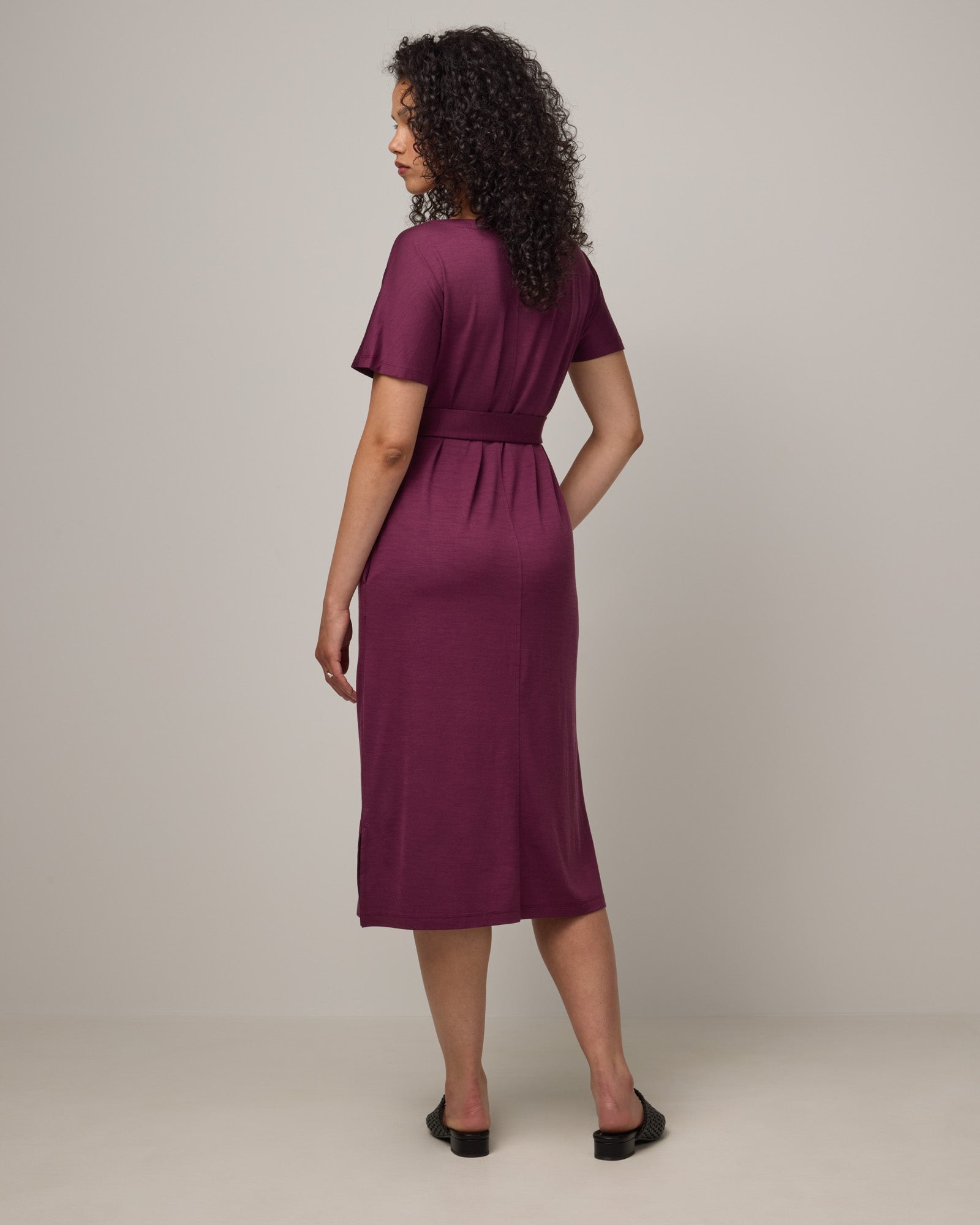 Model wearing Cora Shift Dress in Plum, a reddish-purple color. Cora Shift Dress is a merino wool short sleeve, midi shift dress with pockets and removable belt available in sizes XS-3X.