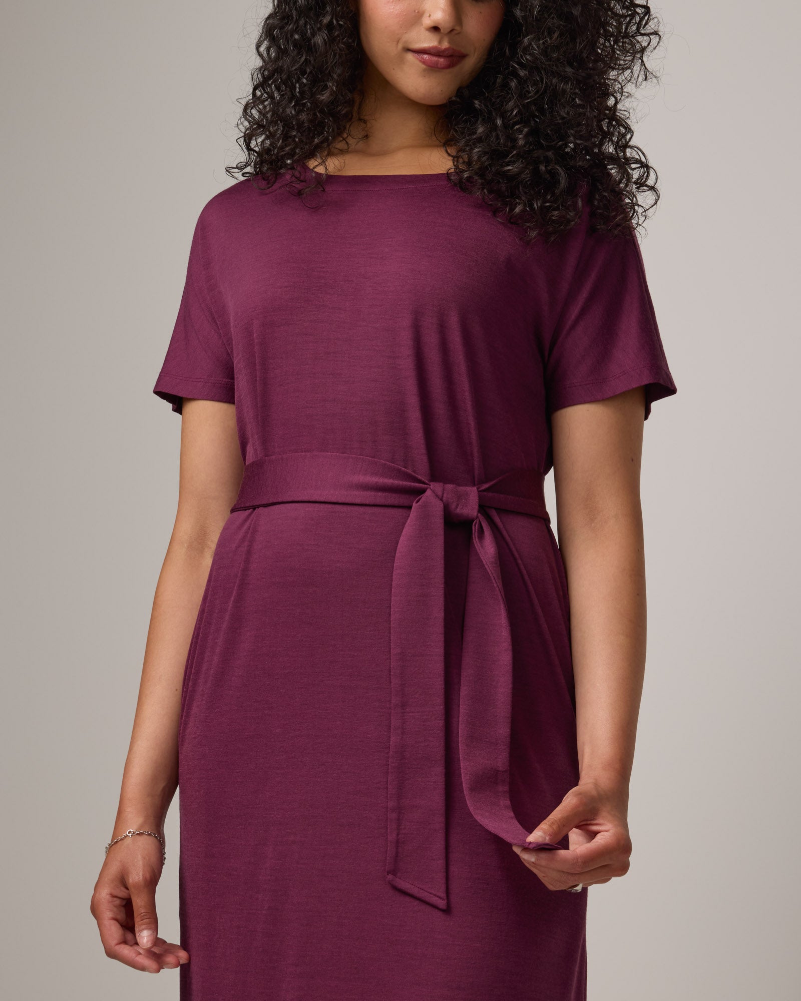 Model wearing Cora Shift Dress in Plum, a reddish-purple color. Cora Shift Dress is a merino wool short sleeve, midi shift dress with pockets and removable belt available in sizes XS-3X.