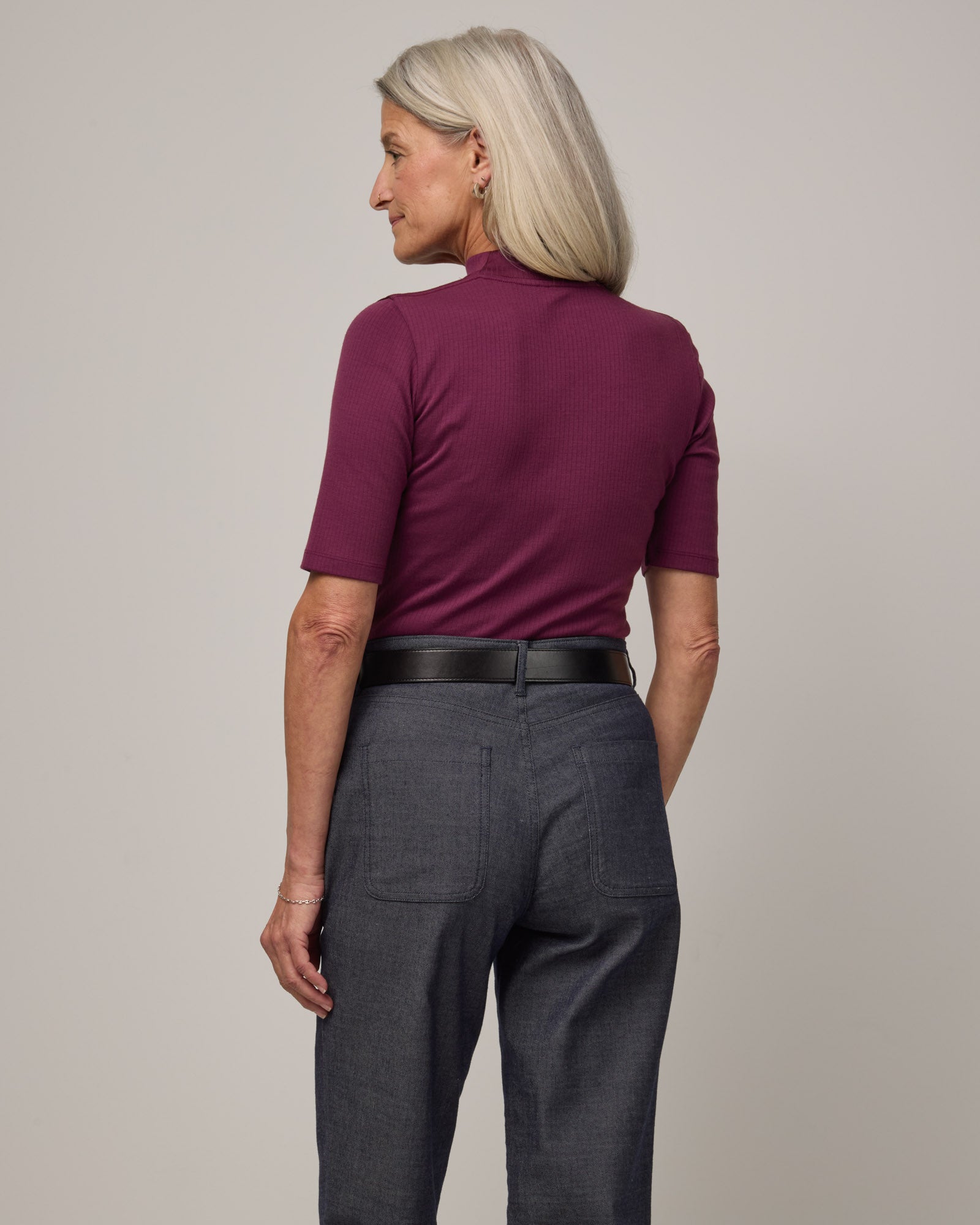 Model wearing Krystal Mock Neck Top in Plum, a reddish-purple color. Krystal Mock Neck Top is a merino wool half-sleeve, mock-neck ribbed tee available in sizes XS-XXL.