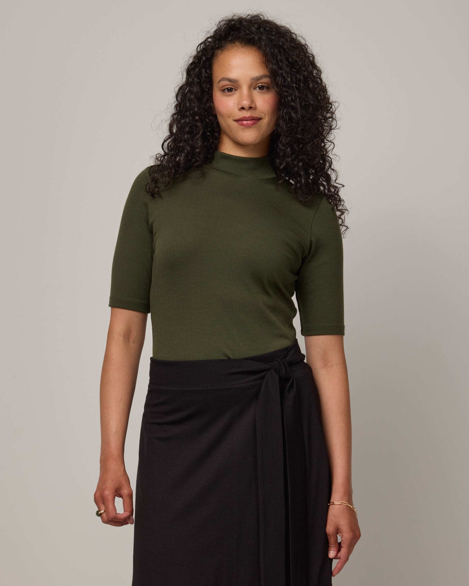 Model wearing Krystal Mock Neck Top in Forest Night, a dark forest green color. Krystal Mock Neck Top is a merino wool half-sleeve, mock-neck ribbed tee available in sizes XS-XXL.