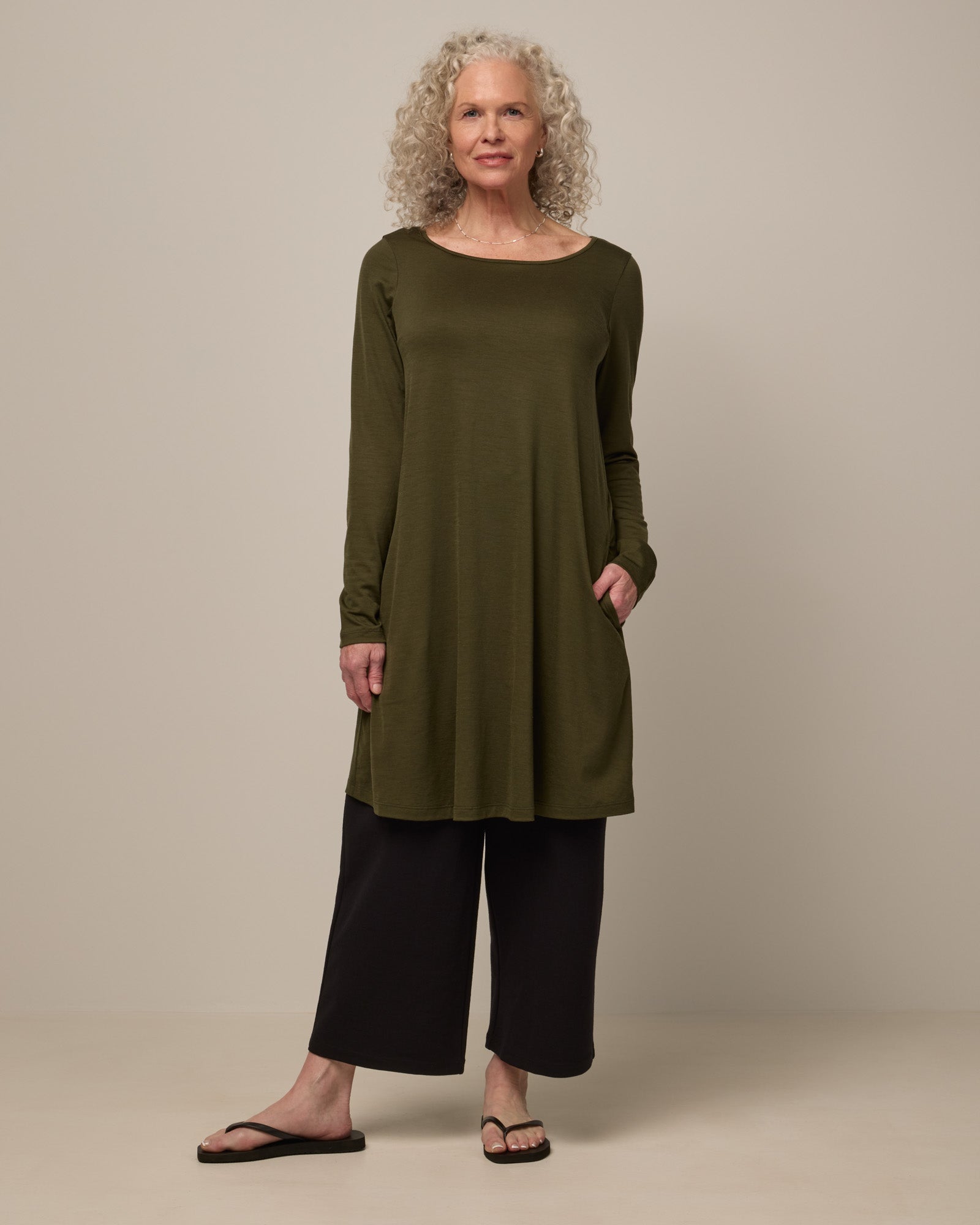 Model wearing Rowena Swing Dress in Forest Night, a dark forest green color. Rowena Swing Dress is a merino wool long sleeve, boatneck, knee length swing dress with pockets available in sizes XS-3X Regular, XS-3X Long.