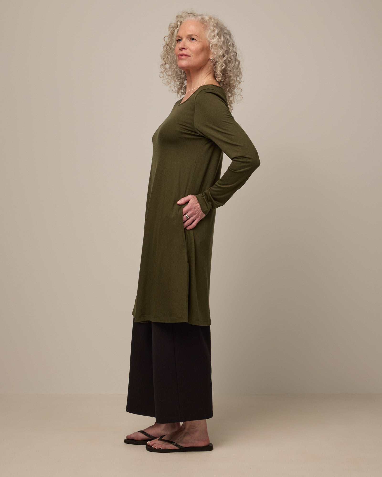 Model wearing Rowena Swing Dress in Forest Night, a dark forest green color. Rowena Swing Dress is a merino wool long sleeve, boatneck, knee length swing dress with pockets available in sizes XS-3X Regular, XS-3X Long.