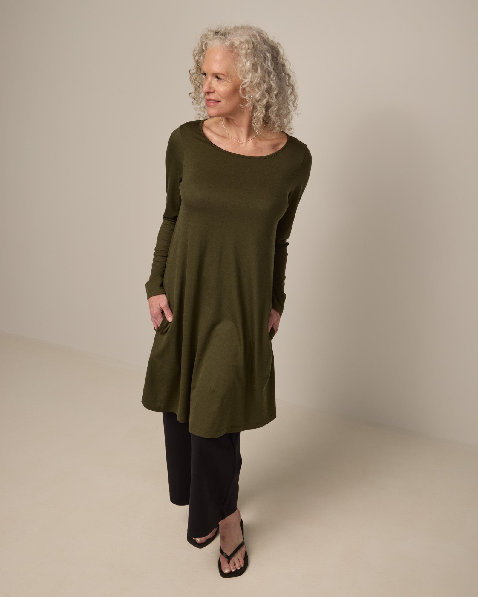 Model wearing Rowena Swing Dress in Forest Night, a dark forest green color. Rowena Swing Dress is a merino wool long sleeve, boatneck, knee length swing dress with pockets available in sizes XS-3X Regular, XS-3X Long.