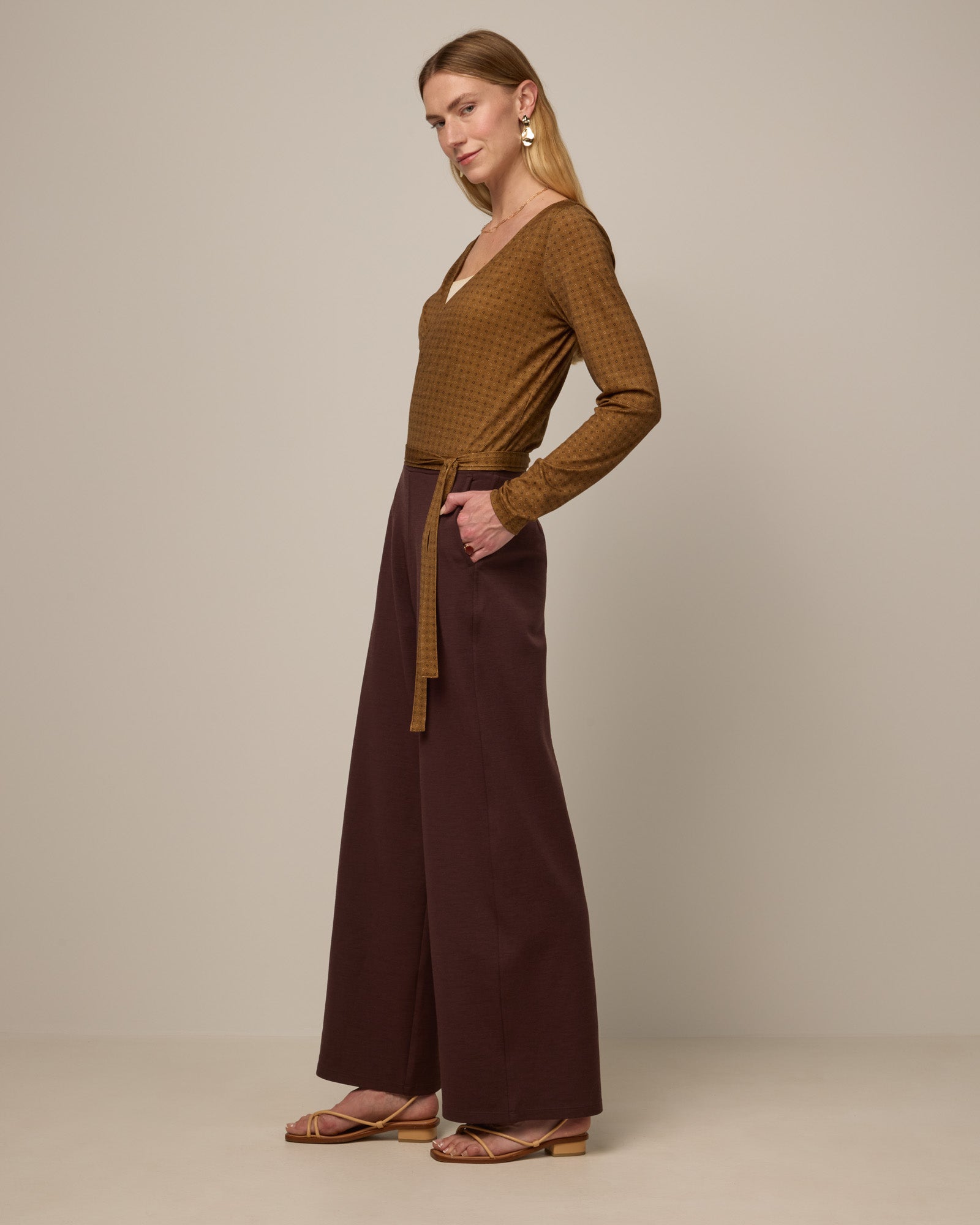 Model wearing Natalia Wrap Top in Geo Stamp Print, a dainty, neutral, geometric print on a dark ochre background. Natalia Wrap Top is a merino wool long sleeve, v-neck, slim fit top available in sizes XS-3X.