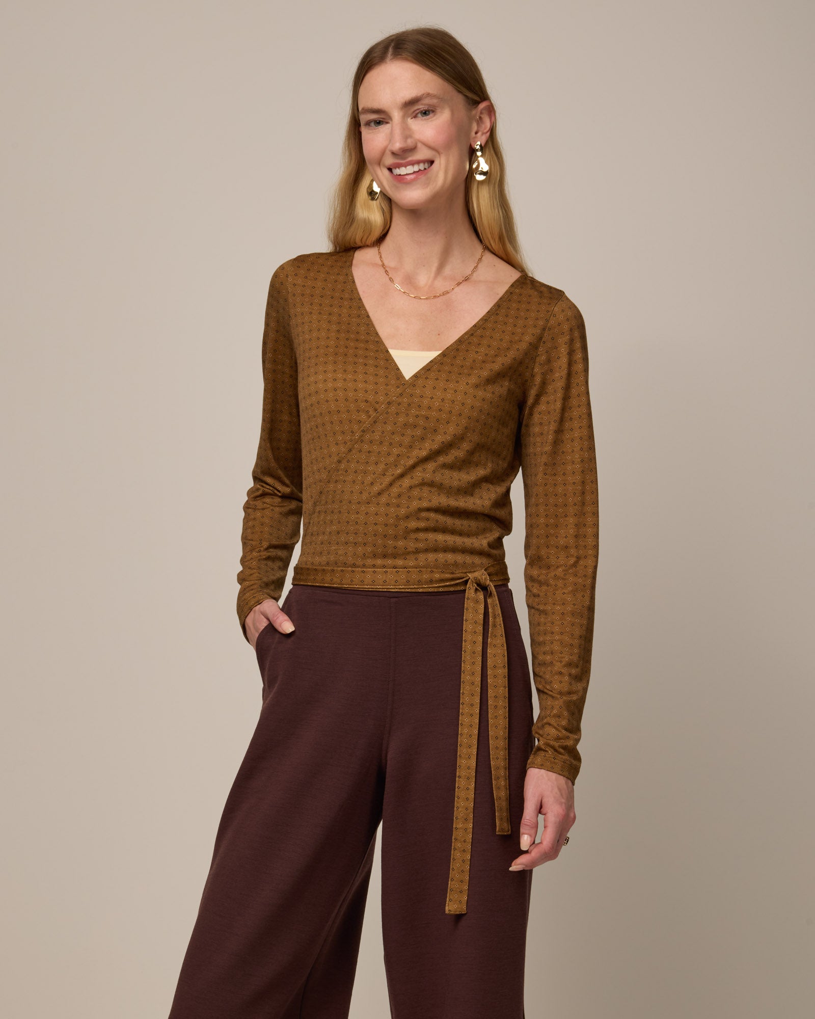 Model wearing Natalia Wrap Top in Geo Stamp Print, a dainty, neutral, geometric print on a dark ochre background. Natalia Wrap Top is a merino wool long sleeve, v-neck, slim fit top available in sizes XS-3X.