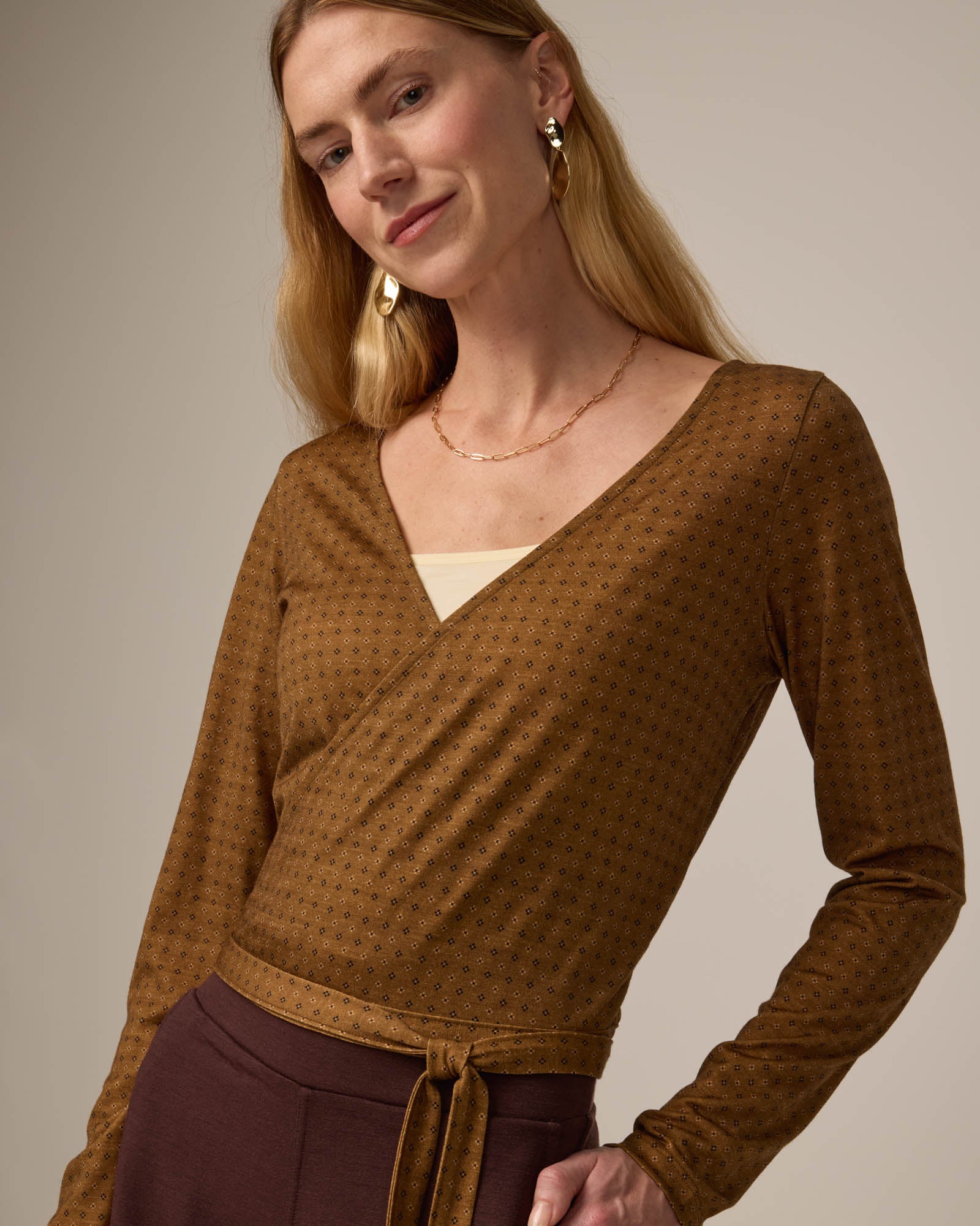 Model wearing Natalia Wrap Top in Geo Stamp Print, a dainty, neutral, geometric print on a dark ochre background. Natalia Wrap Top is a merino wool long sleeve, v-neck, slim fit top available in sizes XS-3X.