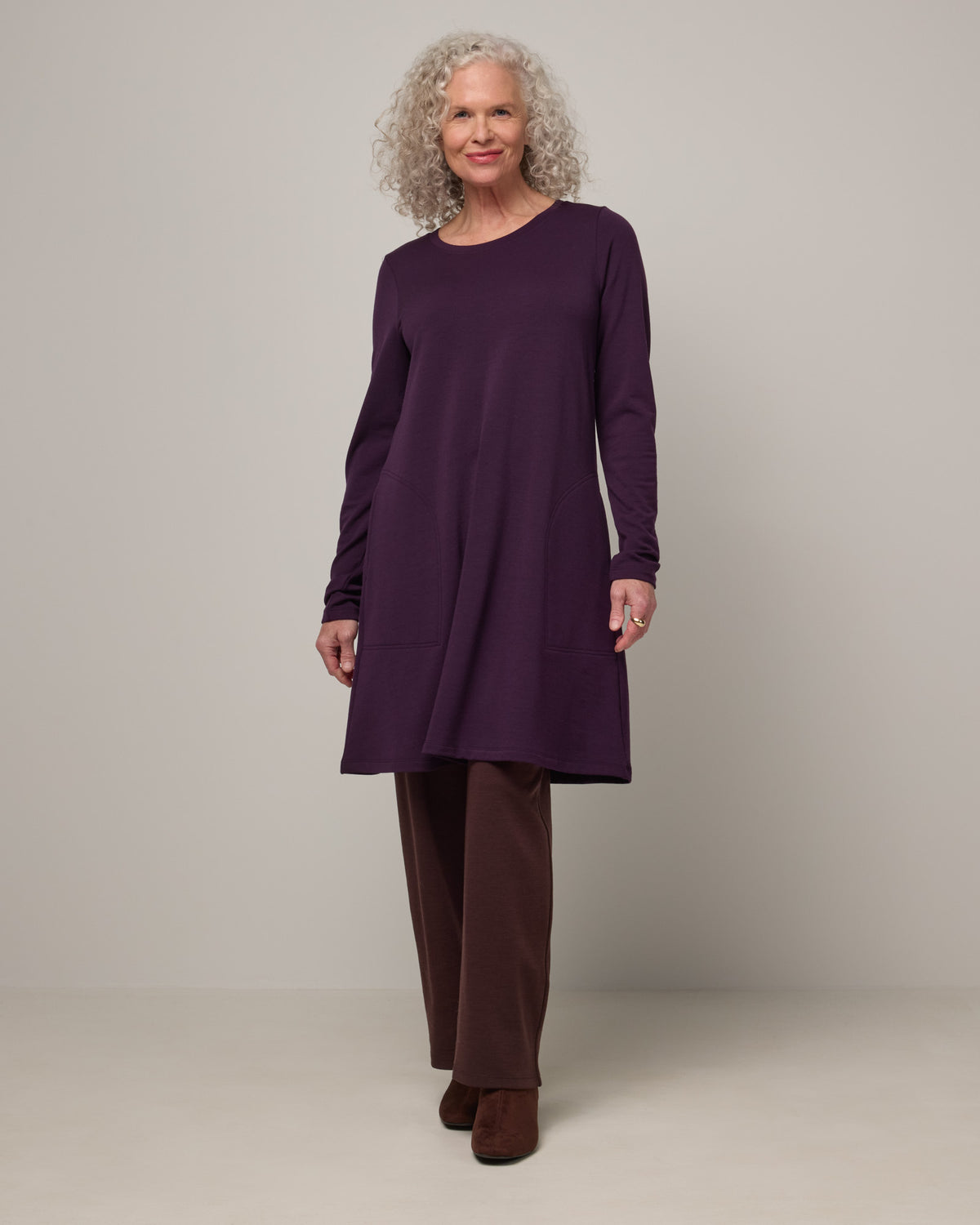 Model wearing Eliza Swing Dress in Cassis, a dark purple color. Eliza Swing Dress is a merino wool long sleeve, crew neck, knee length swing dress with pockets available in sizes XS-3X Regular, XS-3X Long.