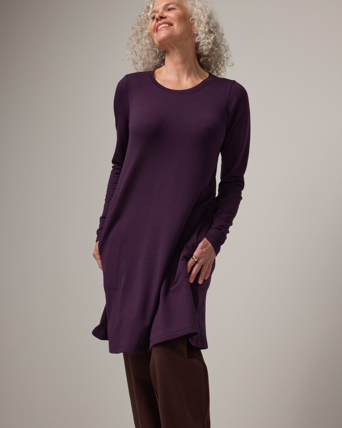 Model wearing Eliza Swing Dress in Cassis, a dark purple color. Eliza Swing Dress is a merino wool long sleeve, crew neck, knee length swing dress with pockets available in sizes XS-3X Regular, XS-3X Long.