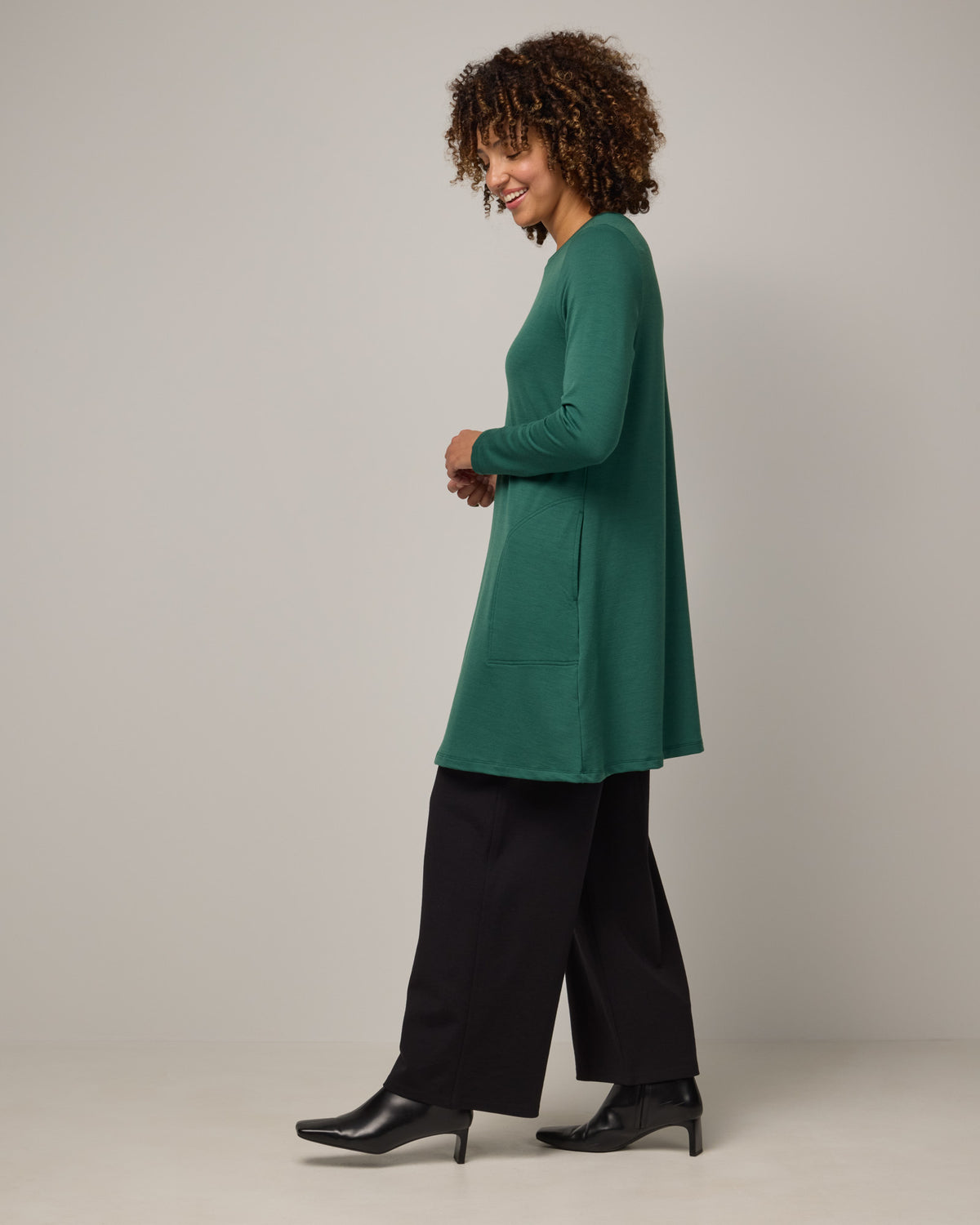Model wearing Eliza Swing Dress in Emerald green. Eliza Swing Dress is a merino wool long sleeve, crew neck, knee length swing dress with pockets available in sizes XS-3X Regular, XS-3X Long.