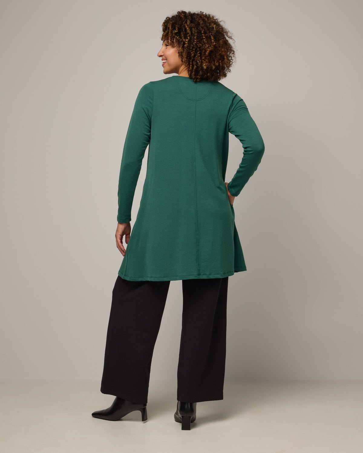 Model wearing Eliza Swing Dress in Emerald green. Eliza Swing Dress is a merino wool long sleeve, crew neck, knee length swing dress with pockets available in sizes XS-3X Regular, XS-3X Long.