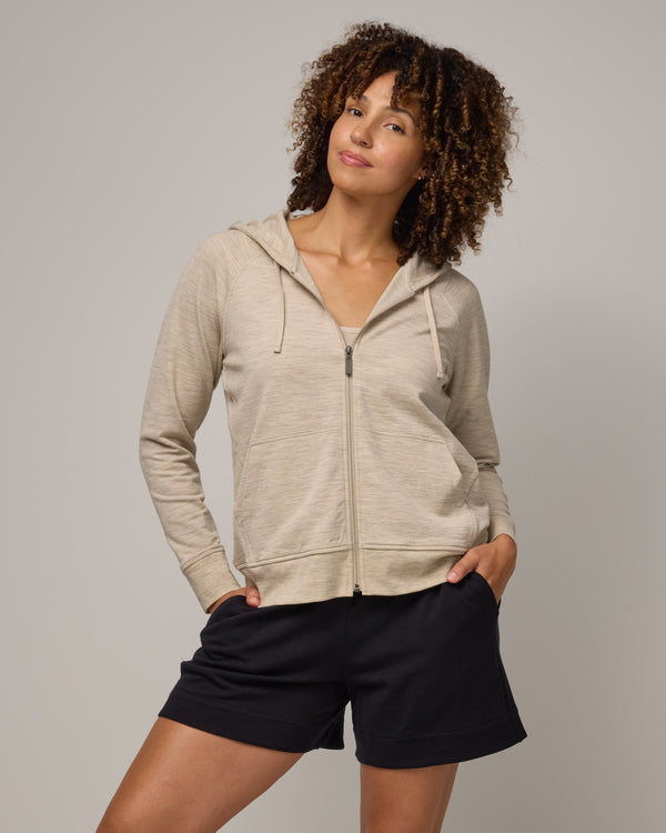Model wearing Aspen Full-Zip Hoodie in Oatmeal Heather, a cream heather color. Aspen Full-Zip Hoodie is a merino wool long sleeve zip hoodie with pockets available in sizes XS-3X.